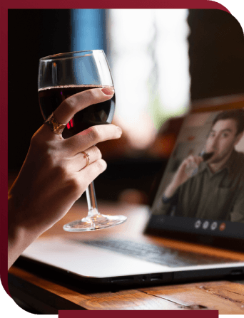 A hand holding a glass of wine, in the background an open laptop displaying another person tasting wine
